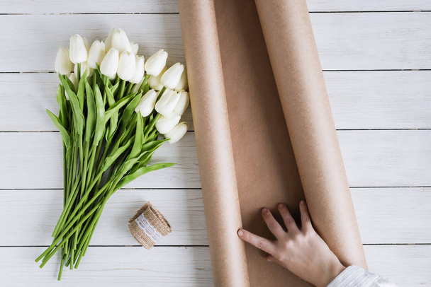 Woman florist wrapping beautiful bouquet of white tulips in pack craft paper on the wooden table. Flat lay - Foto, Bild