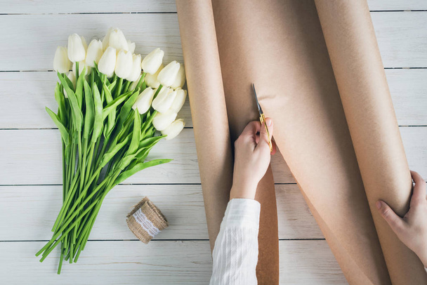 Woman florist wrapping beautiful bouquet of white tulips in pack craft paper on the wooden table. Flat lay - Zdjęcie, obraz
