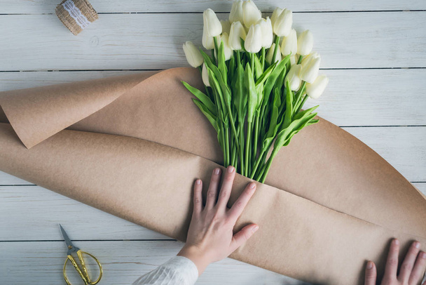 Woman florist wrapping beautiful bouquet of white tulips in pack craft paper on the wooden table. Flat lay - Photo, Image