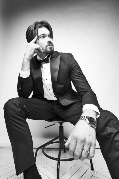 black and white portrait of a handsome young beard man, sitting on the chair, he is wearing  black tuxedo, a white classic shirt with a black bow tie and looking away - Foto, Imagem