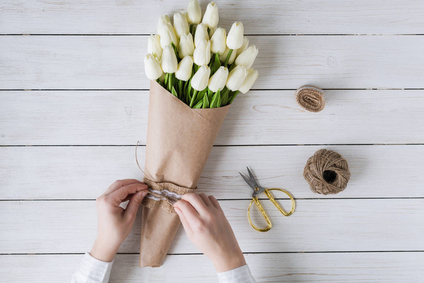 Woman florist wrapping beautiful bouquet of white tulips in pack craft paper on the wooden table. Flat lay - Фото, изображение