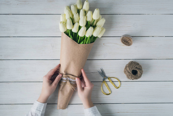 Woman florist wrapping beautiful bouquet of white tulips in pack craft paper on the wooden table. Flat lay - Fotó, kép