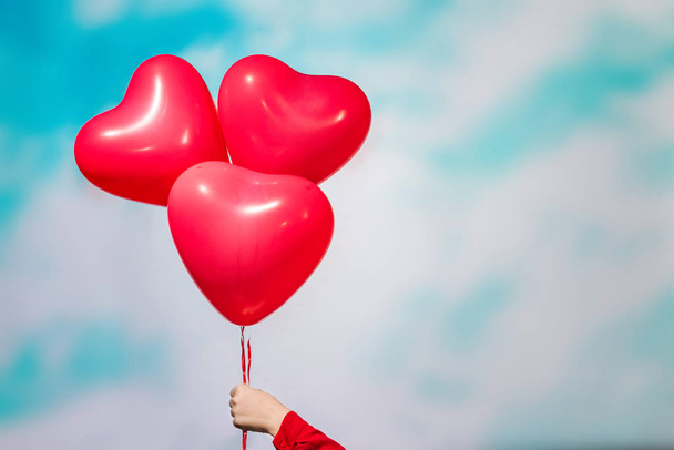  woman hand holding three red balloons in form of heart on the sky background in the blue pastel color. The concept of Valentines day. - Fotó, kép