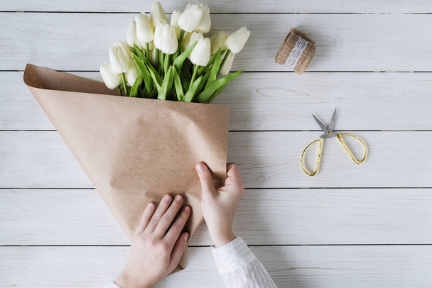 Женщина-флористка заворачивает красивый букет белых тюльпанов в паковую бумагу на деревянный стол. Плоский лежал
 - Фото, изображение