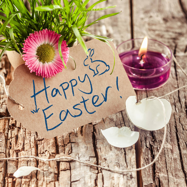 Joyeux vœux de Pâques manuscrits ou salutation sur une étiquette cadeau marron avec une coquille d'oeuf cassée, herbe fraîche avec une fleur de printemps colorée et une bougie allumée, fond en bois rustique
 - Photo, image