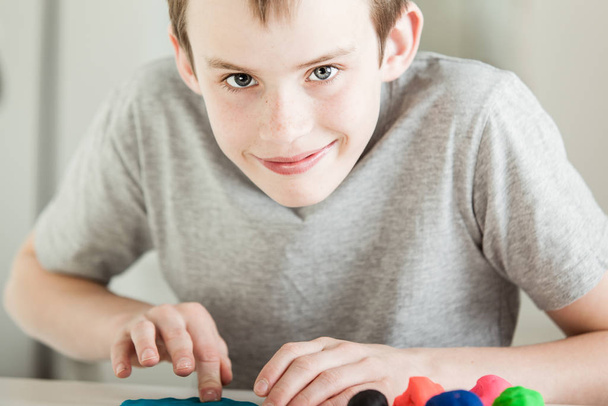Single happy boy with grin in gray shirt pressing into clay pieces of various colors with fingers on table - Fotoğraf, Görsel