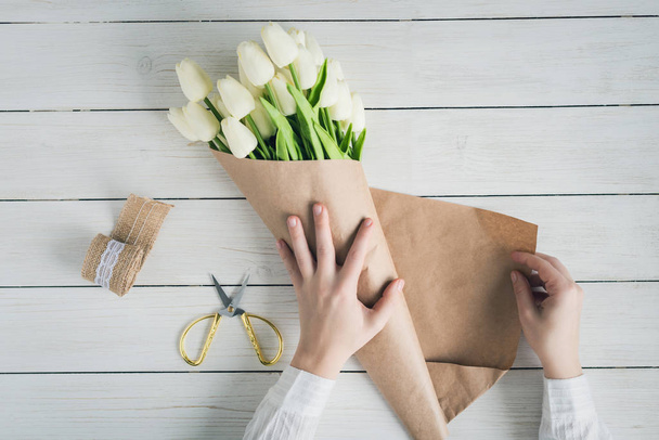 Woman florist wrapping beautiful bouquet of white tulips in pack craft paper on the wooden table. Flat lay - Foto, Imagen