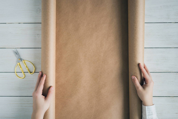 Female hands unfolding a roll of wrapping brown craft paper for packing gifts and flowers on the wooden white table. Copy space.  - Zdjęcie, obraz