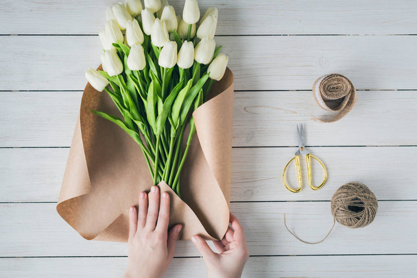 Woman florist wrapping beautiful bouquet of white tulips in pack craft paper on the wooden table. Flat lay - Фото, зображення