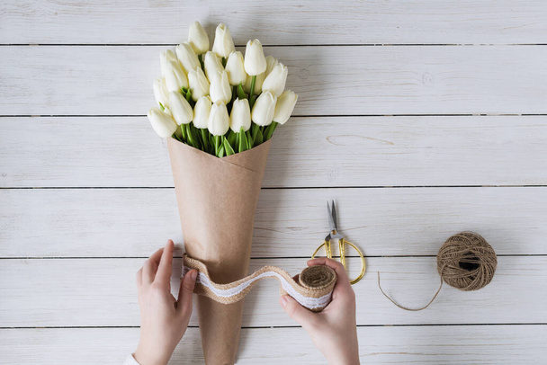 Woman florist wrapping beautiful bouquet of white tulips in pack craft paper on the wooden table. Flat lay - 写真・画像