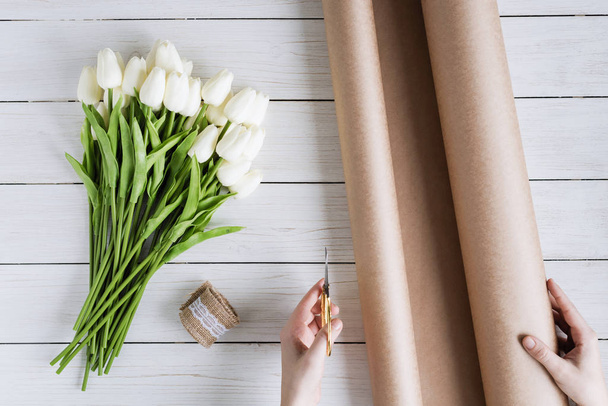 Woman florist wrapping beautiful bouquet of white tulips in pack craft paper on the wooden table. Flat lay - Foto, afbeelding
