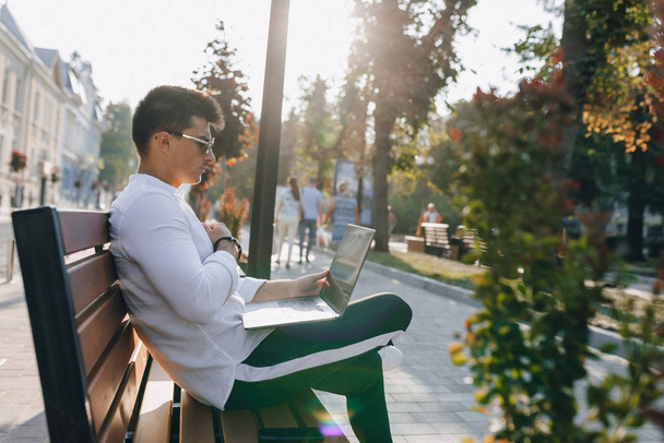 молодий стильний хлопець в сорочці з телефоном і блокнотом на лавці в сонячний теплий день на відкритому повітрі, фрілансер
 - Фото, зображення