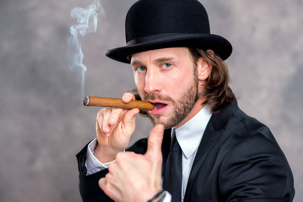 young businessman with bowler hat and thumb up smoking big cigar - Фото, изображение