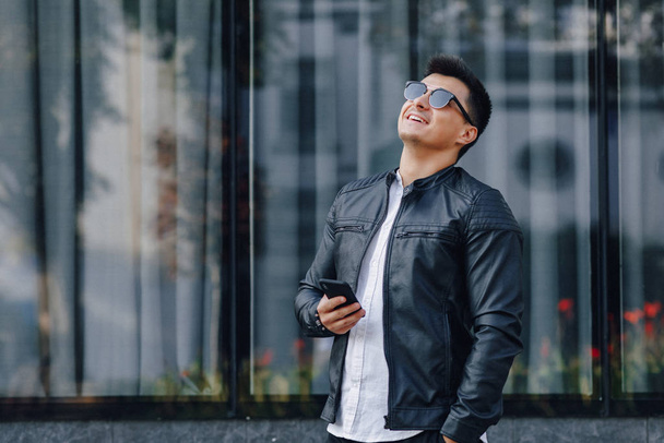young stylish guy in glasses in black leather jacket with phone on glass background - Zdjęcie, obraz