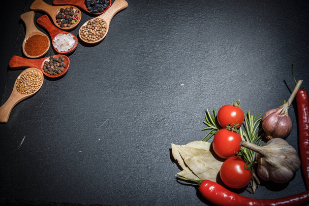 Spices in wooden spoons and cherry tomatoes, chili peppers, garlic and herbs for cooking on a black surface and black background - Zdjęcie, obraz