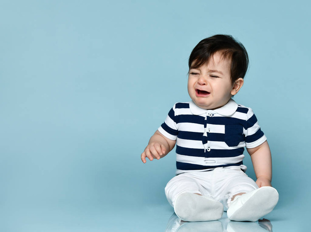 Little kid in striped t-shirt, white pants and booties. He is about to cry, sitting on floor against blue background. Close up - Photo, Image