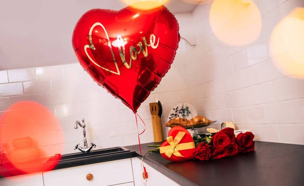 Little presents. Close-up photo of a big heart-shaped balloon, a bouquet of roses and a red heart-shaped box with a golden ribbon, lying on a table in the kitchen. - Фото, изображение