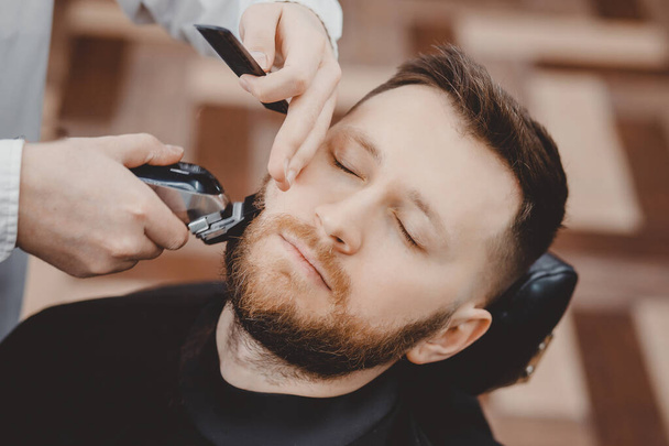Holič stříhá vousy chlapovi v holičství, zarámuje vlasy. elektrický holicí strojek, retro tónovaná hnědá - Fotografie, Obrázek