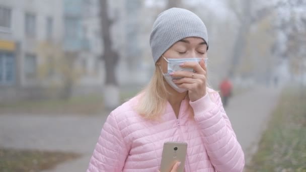 Coronavirusbescherming. Een jonge vrouw met een medisch beschermend masker staat met een smartphone op een straat in Europa. Symptomen van het coronavirus uit China kwamen naar voren. - Video