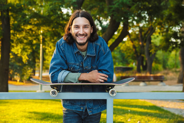 Smiling positive optimistic young bearded man with skateboard standing in nature park. - Φωτογραφία, εικόνα