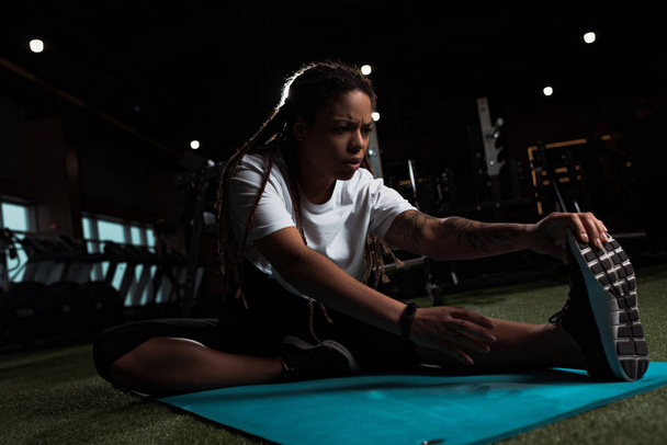 African american woman sitting and stretching on fitness mat in gym - Foto, immagini