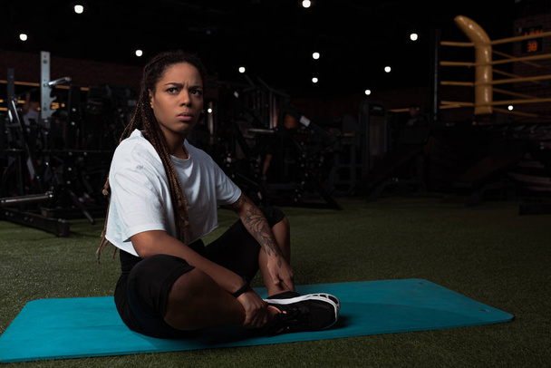 Worried and displeased african american woman sitting with crossed legs on fitness mat - Photo, Image