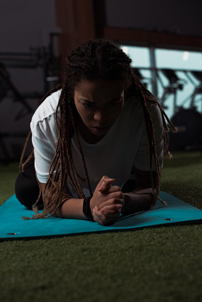 Estressado e desagradado afro-americano mulher sentada com as mãos apertadas no tapete de fitness
  - Foto, Imagem