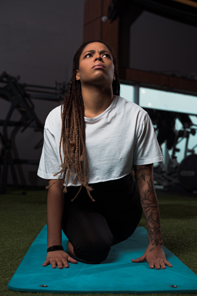 Thoughtful and worried african american woman sitting on fitness mat in gym - Fotoğraf, Görsel