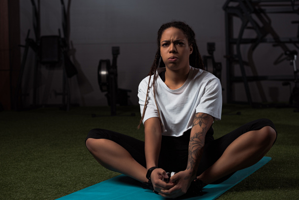  Femme afro-américaine tendue assise avec les mains serrées sur le tapis de fitness
  - Photo, image