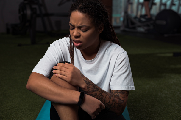 Worried and upset african american woman suffering from pain in knee, sitting on fitness mat - Photo, Image