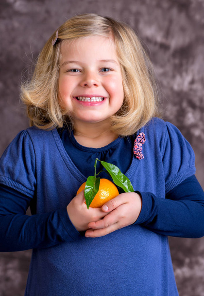 little funny girl with mandarin orange in front of brown background - Foto, Imagem