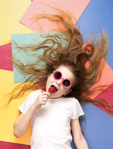 Vibrant colors: a child girl lies on a colorful background and licks a lollipop. - Foto, imagen