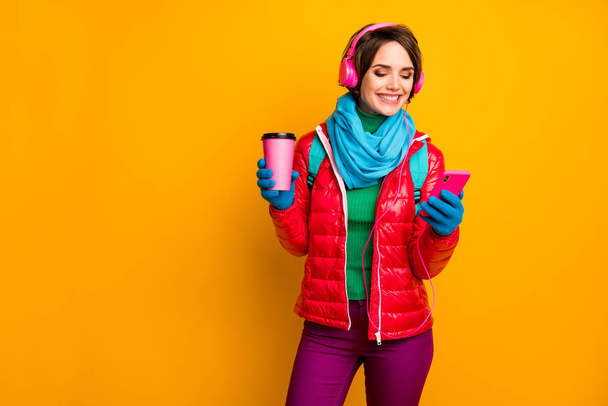 Photo of funny blogger lady walk street hold telephone listening earphones youth music drink hot coffee wear casual red coat blue scarf gloves pants isolated yellow color background - 写真・画像