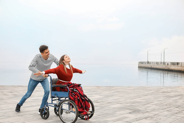 Инвалидная молодая женщина в инвалидной коляске и ее муж возле реки
 - Фото, изображение