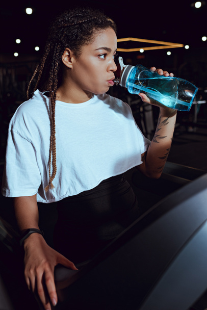 Enfoque selectivo de la mujer afroamericana bebiendo agua de la botella deportiva en la cinta de correr
 - Foto, Imagen