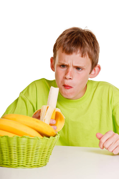 Unimpressed young boy eating a fresh banana with a look of horror on his face as he chews the first mouthful while seated at the table - Photo, Image