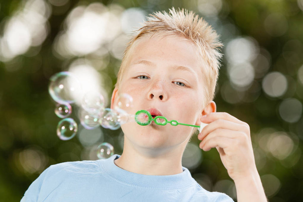 Close up of cute little blond boy with spiky hair and blue shirt blowing bubbles from green wand outdoors - Fotografie, Obrázek