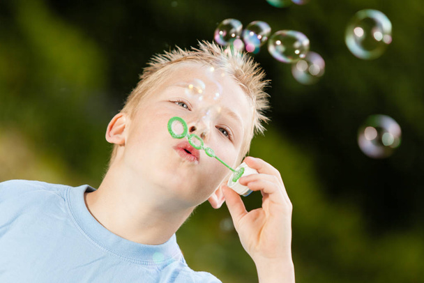 Close up on happy little blond boy with spiked hair and blue shirt blowing bubbles upward from green wand outdoors - Photo, Image
