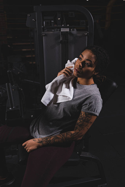 tatuado afro-americano menina segurando toalha perto de máquina de fitness
 - Foto, Imagem