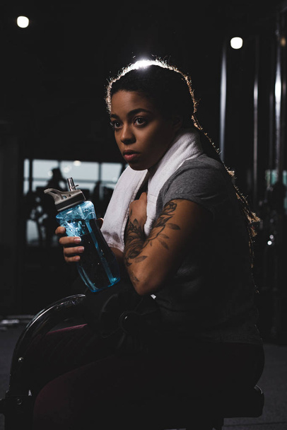 νεαρή Αφροαμερικανή με τατουάζ που κρατά αθλητικό μπουκάλι στο γυμναστήριο  - Φωτογραφία, εικόνα