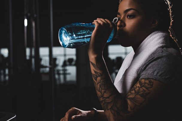 Αφροαμερικανή κοπέλα με τατουάζ πόσιμο νερό, ενώ κρατώντας αθλητικό μπουκάλι στο γυμναστήριο  - Φωτογραφία, εικόνα