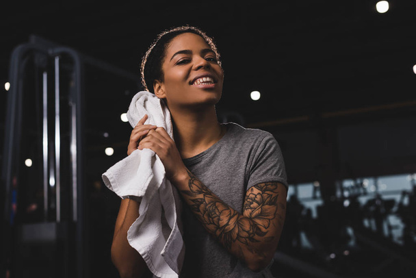 Τατουάζ αφροαμερικανή κοπέλα σκούπισμα ιδρώτα με πετσέτα και χαμογελώντας n γυμναστήριο  - Φωτογραφία, εικόνα