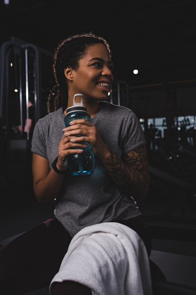 χαρούμενη και με τατουάζ Αφροαμερικανή γυναίκα που κρατάει αθλητικό μπουκάλι  - Φωτογραφία, εικόνα