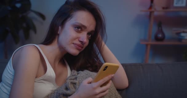 Close up of depressed girl sitting on sofa with smartphone in fetal position . Young unhappy woman with tears on her face looking at mobile phone screen and thinking of bad news. - Záběry, video