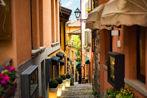 Bellagio kasabasında turuncu binalar ve fenerlerle dolu dar bir sokak ünlü İtalyan gölü Como, Lombardy, Kuzey İtalya 'da taş basamakları ve arka planda Alp dağlarıyla. - Fotoğraf, Görsel