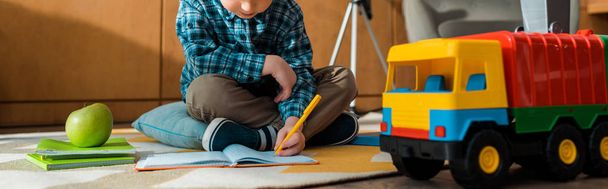 panoramiczne ujęcie inteligentnego dziecka piszącego w notatniku w pobliżu zabawkowego samochodu i jabłka siedząc na podłodze  - Zdjęcie, obraz