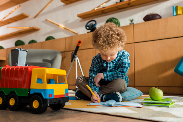 inteligentne dziecko piszące w notatniku siedząc na podłodze w pobliżu jabłka, zabawkowego samochodu i książek  - Zdjęcie, obraz
