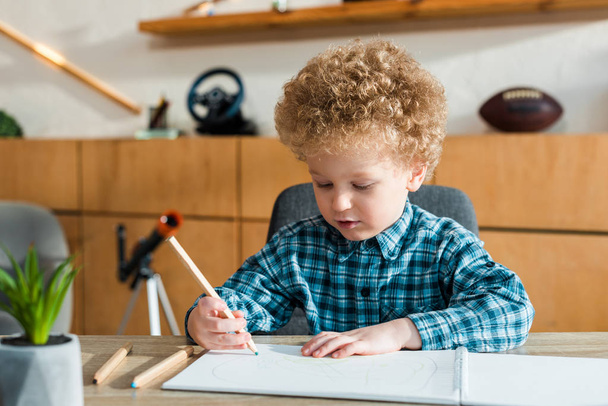 selective focus of smart and curly kid holding pencil while drawing on blank paper  - Фото, изображение
