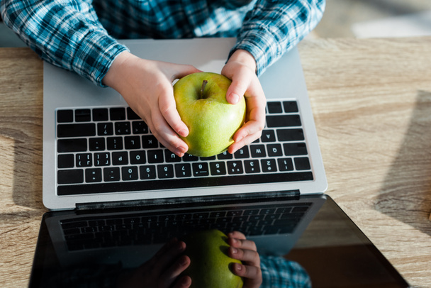 cropped view of kid holding fresh apple near laptop with blank screen  - Photo, Image
