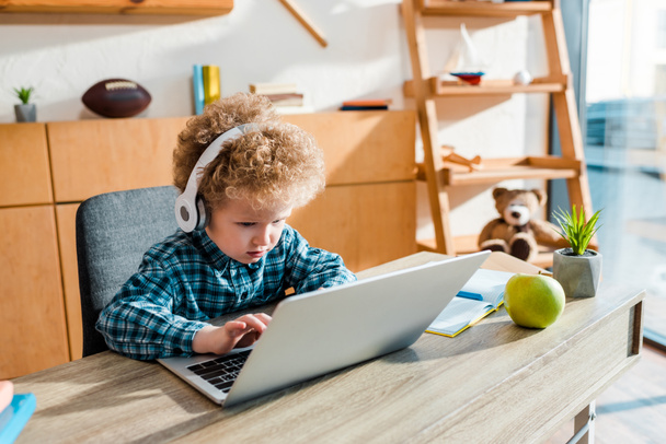 garoto encaracolado digitando no laptop perto da maçã enquanto ouve música em fones de ouvido sem fio
  - Foto, Imagem